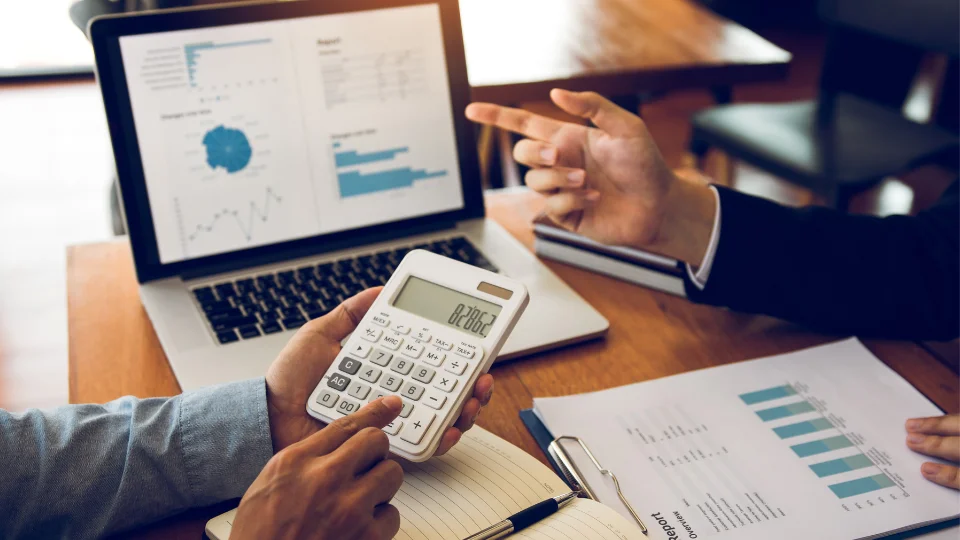 Two business professionals review documents and use a calculator at a table, analyzing the costs of outsourcing payroll services