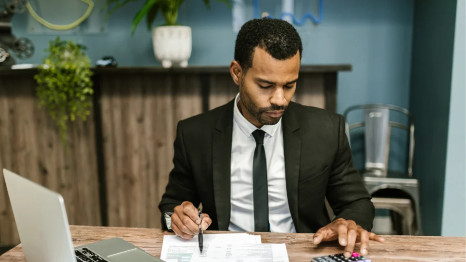 An accounting professional calculates payroll costs using a calculator, with financial documents and a laptop open on the desk.