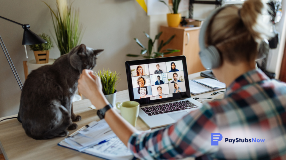 a person sitting at a desk in a home office environment, with a cat nearby and an open laptop displaying a video call
