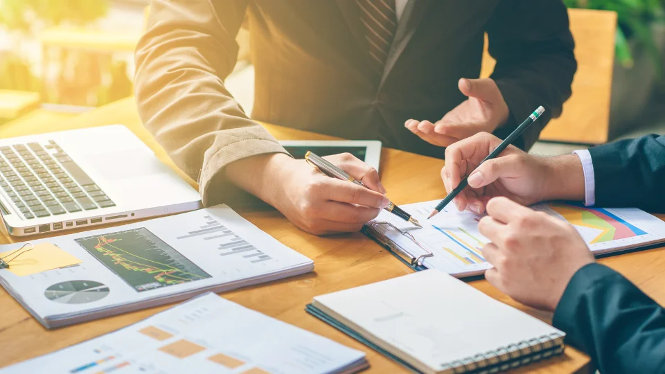 Two people sit at a desk reviewing documents with financial metrics for tracking income and expenses