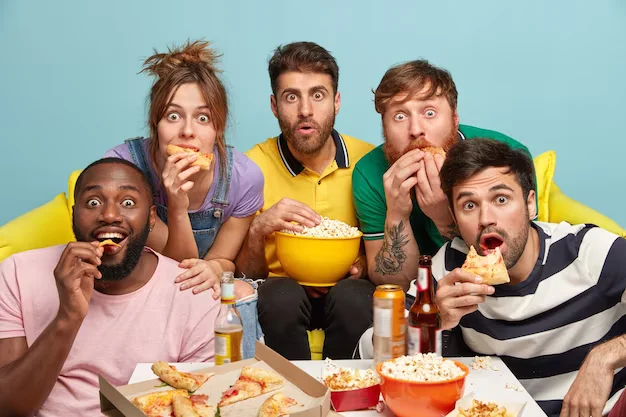 Indoor shot of surprised mixed race friends, eat popcorn, pizza, have fearful panicked faces, enjoy watching horror films, have free time on evening weekend, sit on sofa. People and free time concept